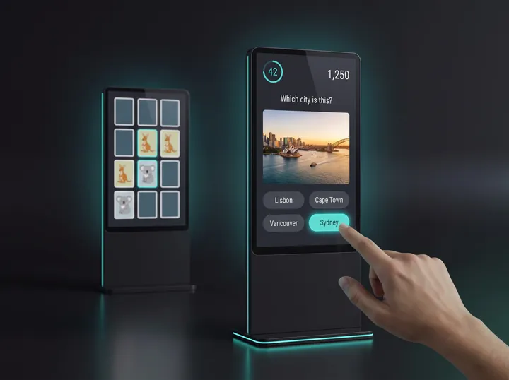 A touchscreen kiosk showing a city quiz with four answers, a finger tapping the right one, and a second kiosk with a memory game beside it